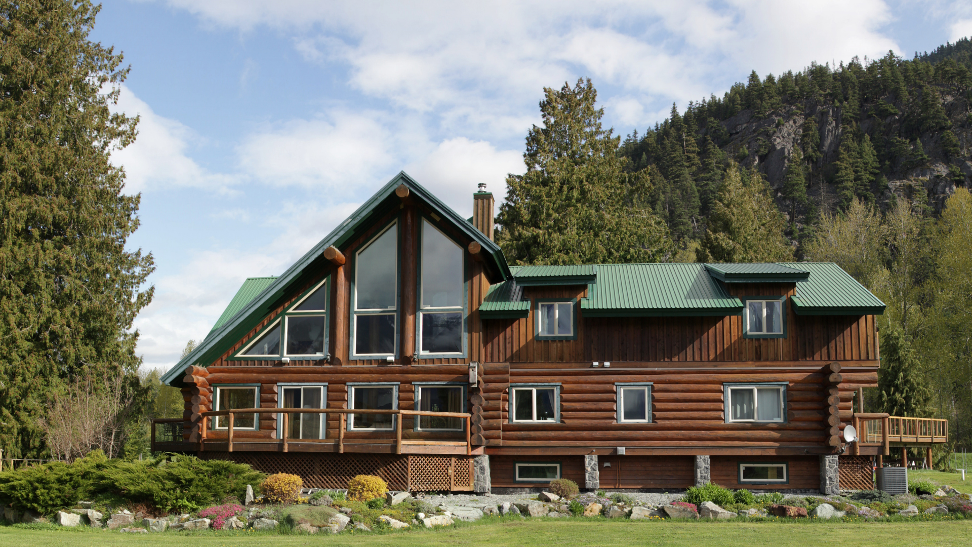 Log cabin house with green roof, large windows, and mountain backdrop.
