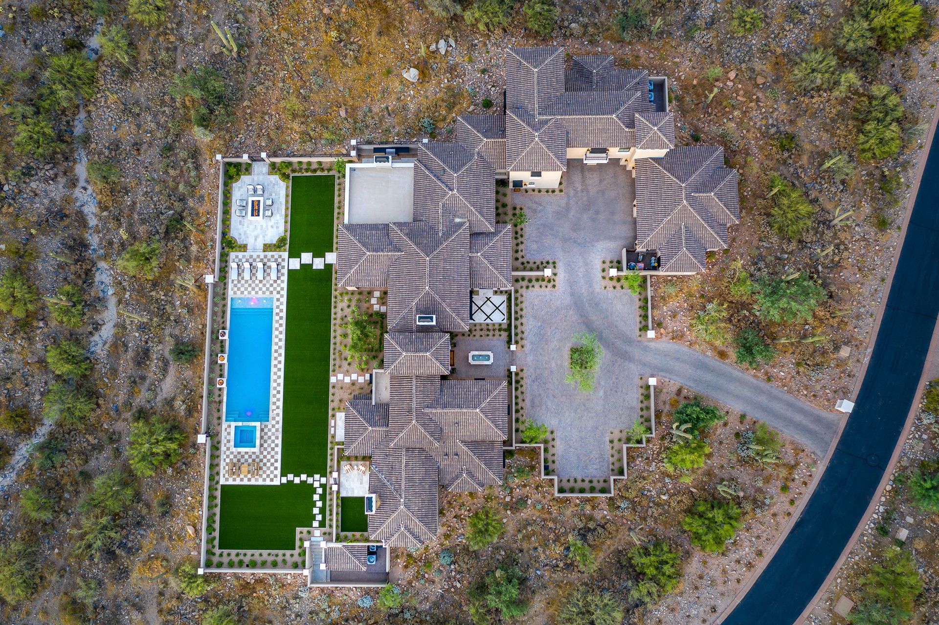 Aerial view of a resort style custom home build with a long blue pool beside a curved road and trees.
