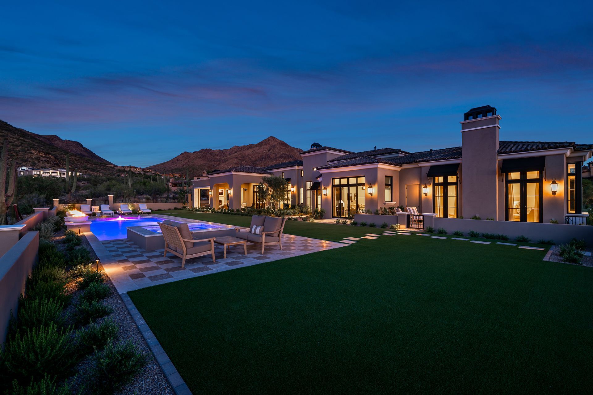 Luxury resort style home at dusk with lit pool, loungers, and modern backyard under a blue sky
