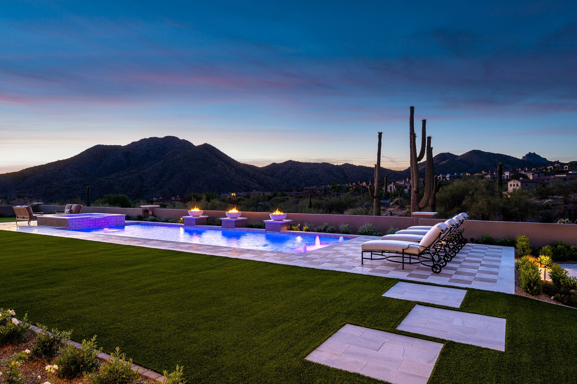Outdoor pool with blue-purple lighting beside a grassy lawn, lounge chairs, and hills at dusk