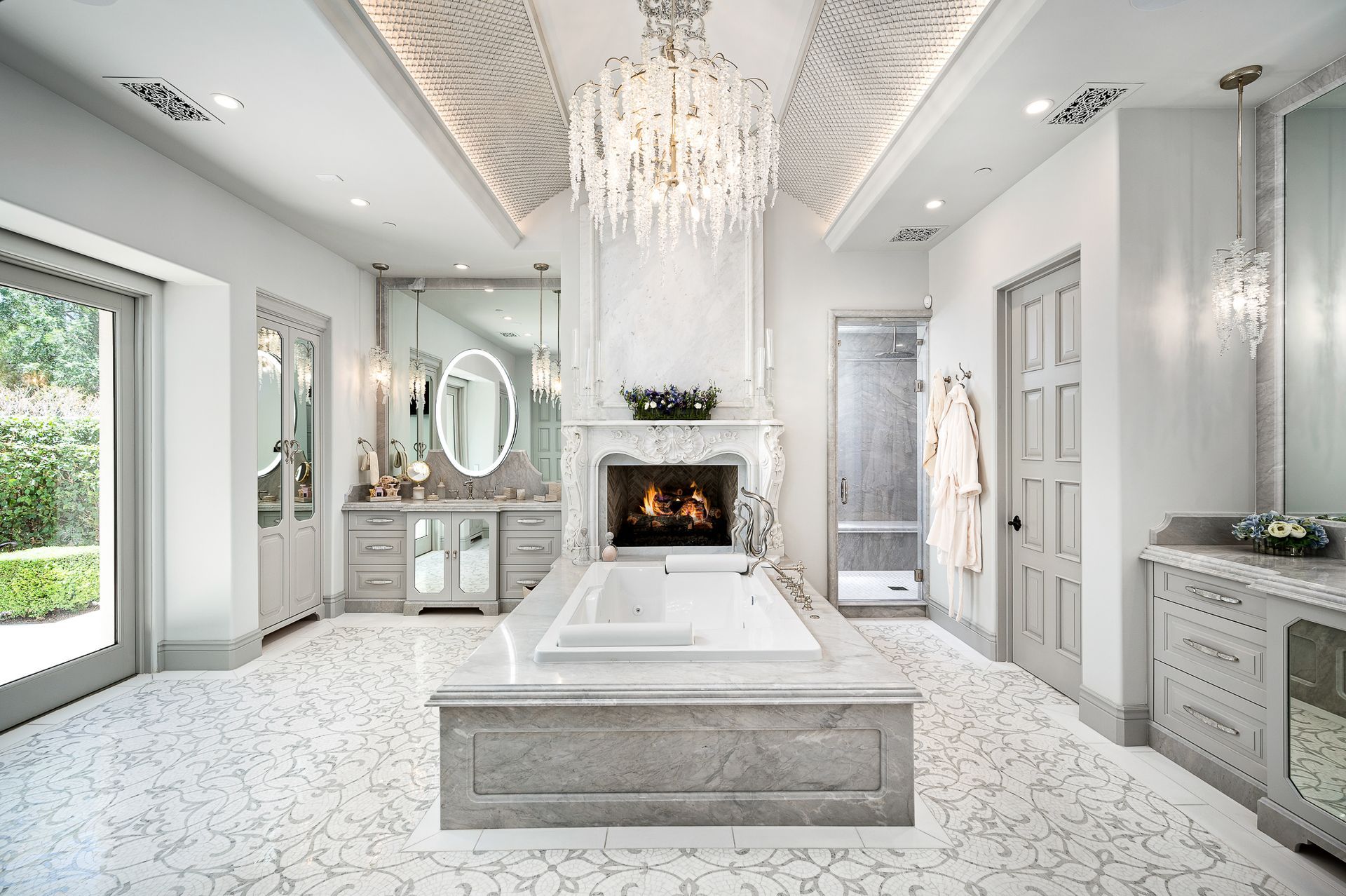 A luxurious, custom white-toned bathroom interior design featuring a central bathtub, fireplace, crystal chandelier, and patterned floor tiling.