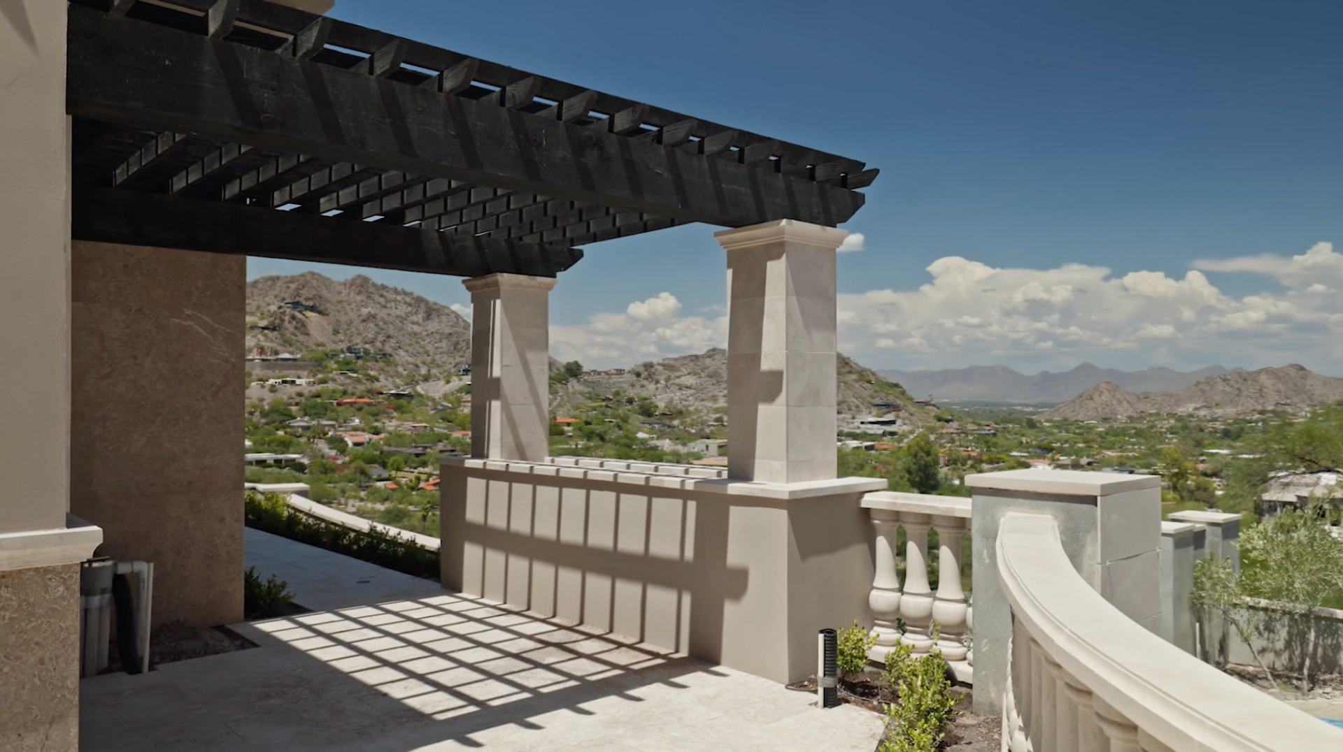 Sunlit balcony with white railings and mountain view under a dark wooden pergola