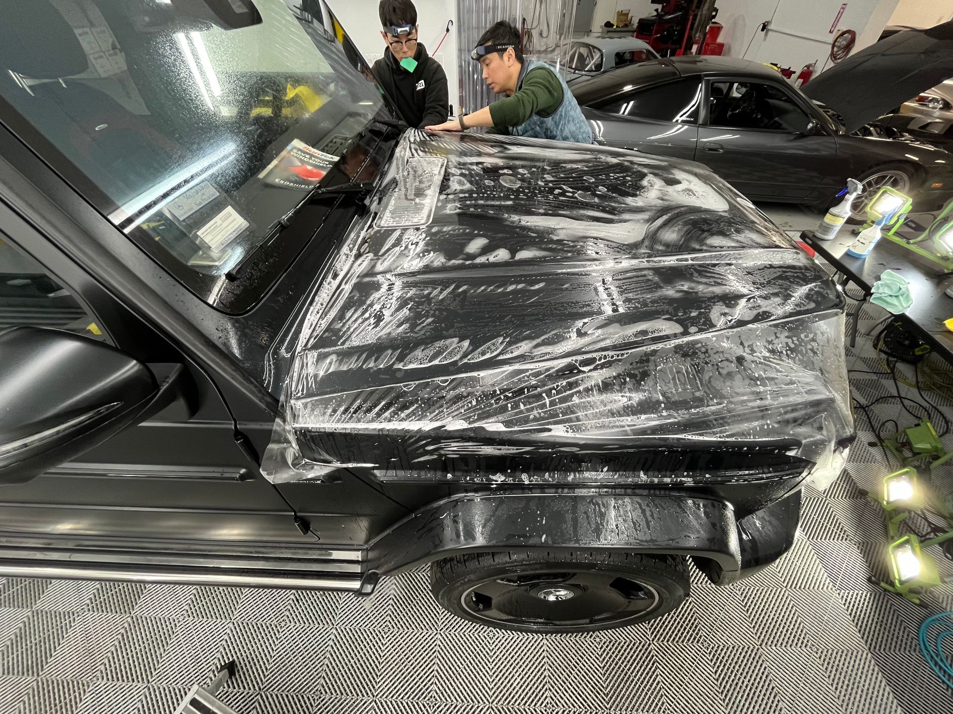 Black car hood wrapped in protective film while two people inspect it in a garage.
