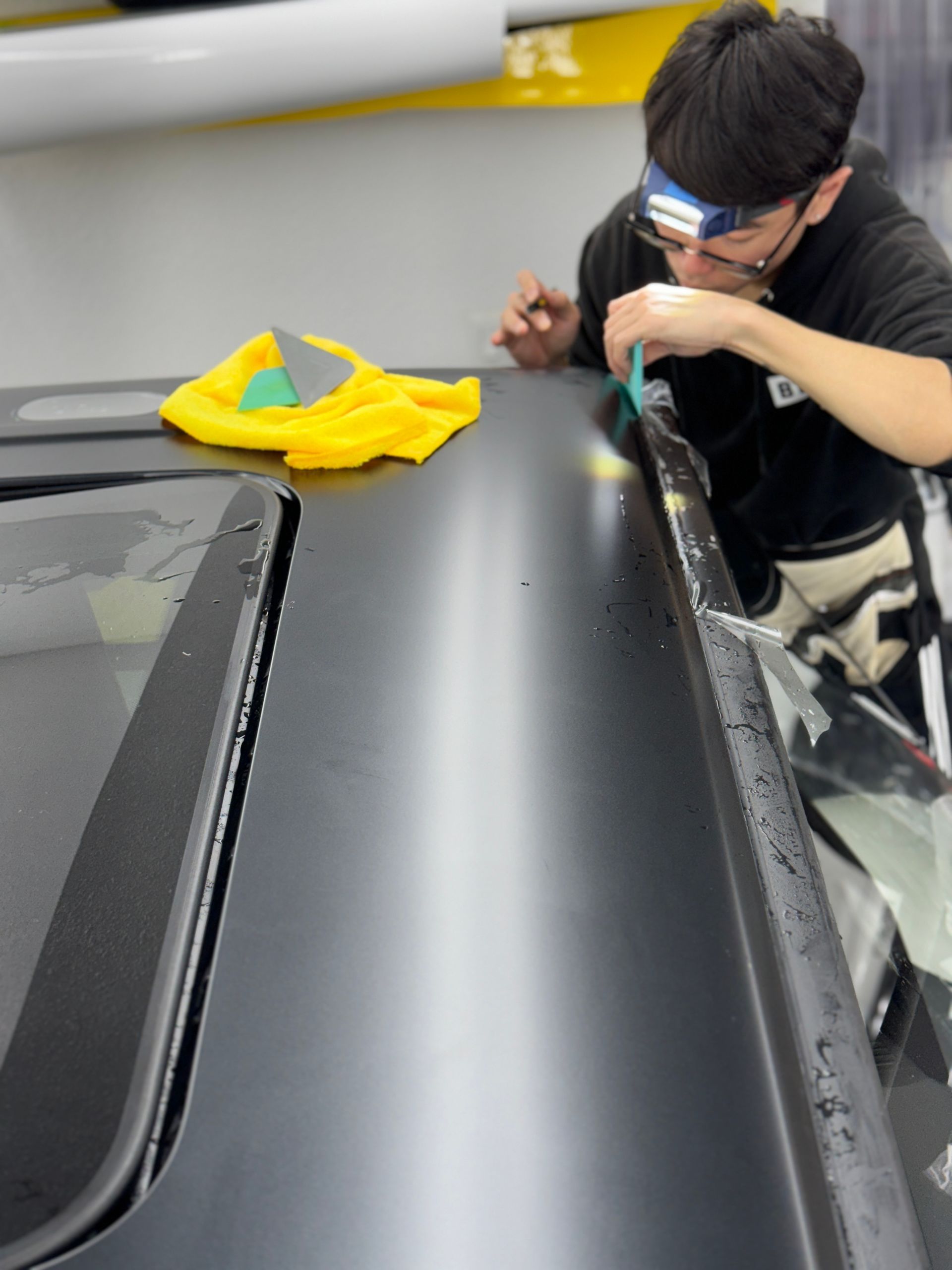Person cleaning a black car roof with a cloth in a garage, with yellow microfiber towels nearby
