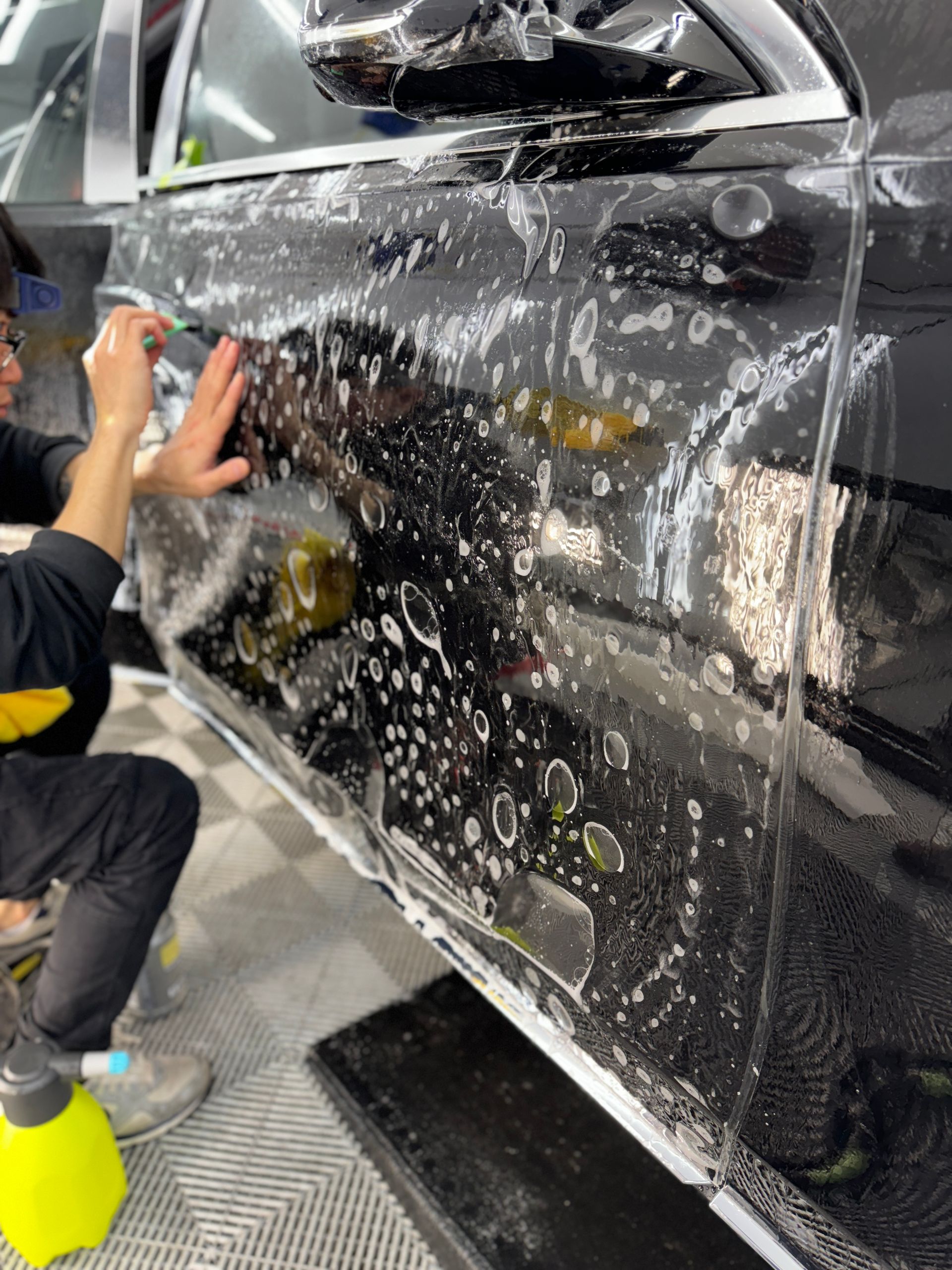 Person washing a black car covered in soap suds in a garage or car wash