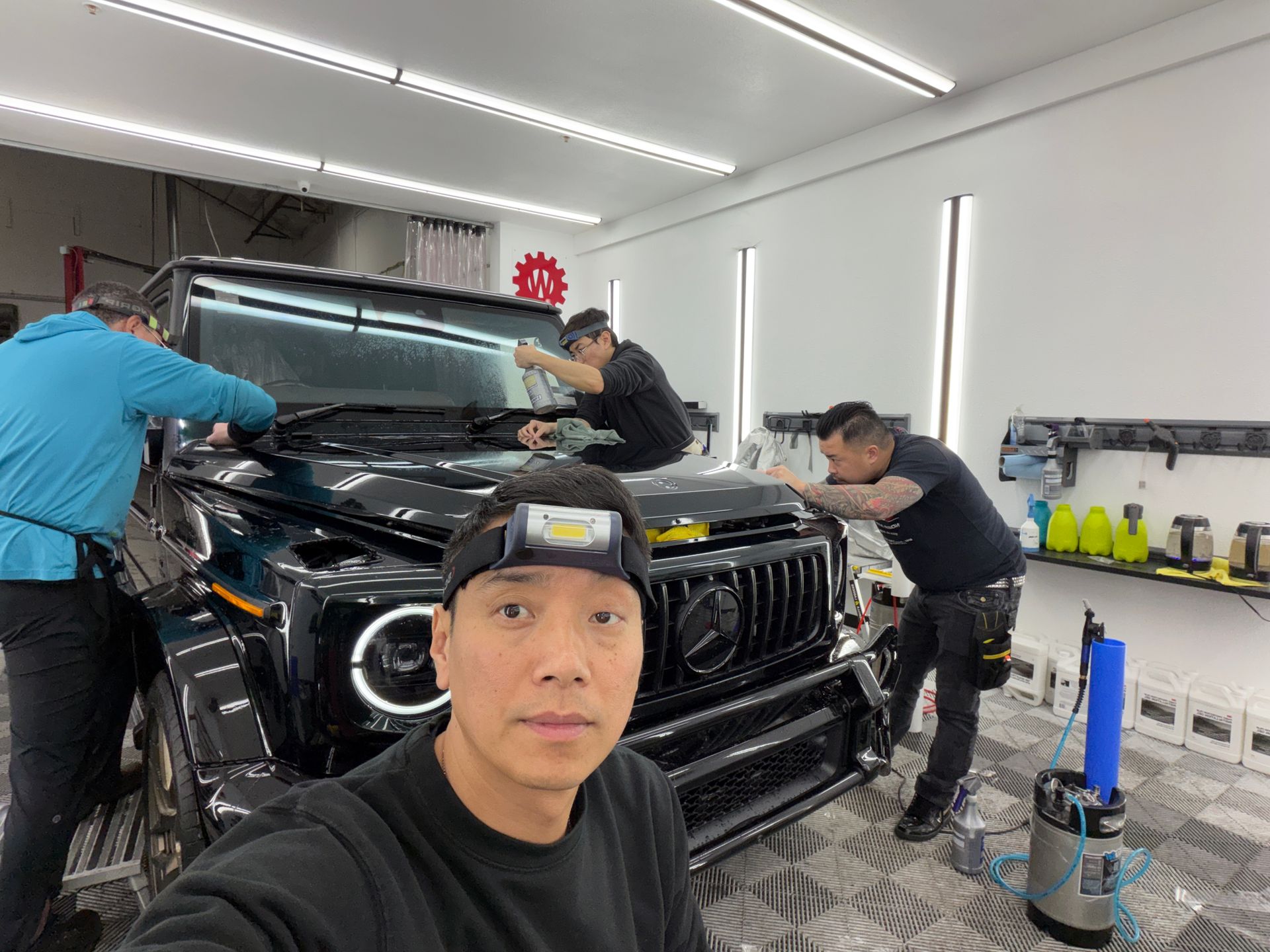 Four people detailing a black SUV in a bright garage, with one person taking a selfie in the foreground.