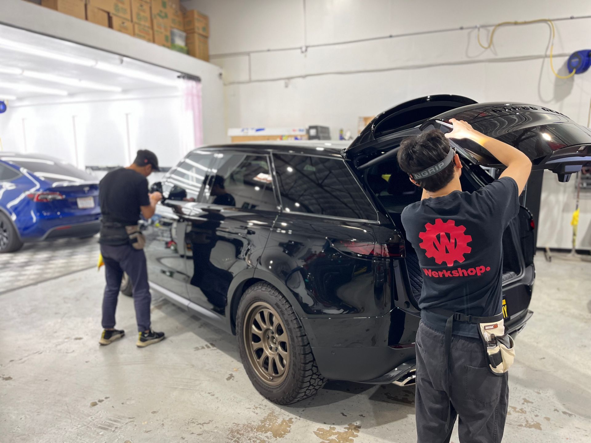 Mechanics working on a black SUV in a brightly lit garage, one worker lifting the open rear hatch