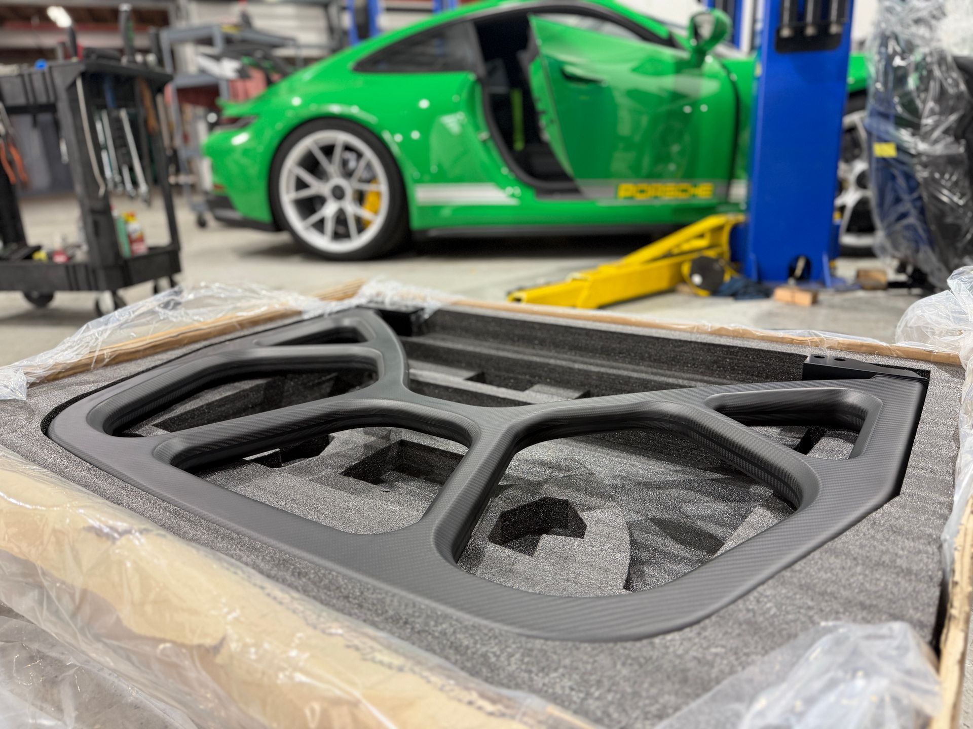Black carbon-fiber automotive grille panel on a garage workbench, with a green sports car and lift in the background