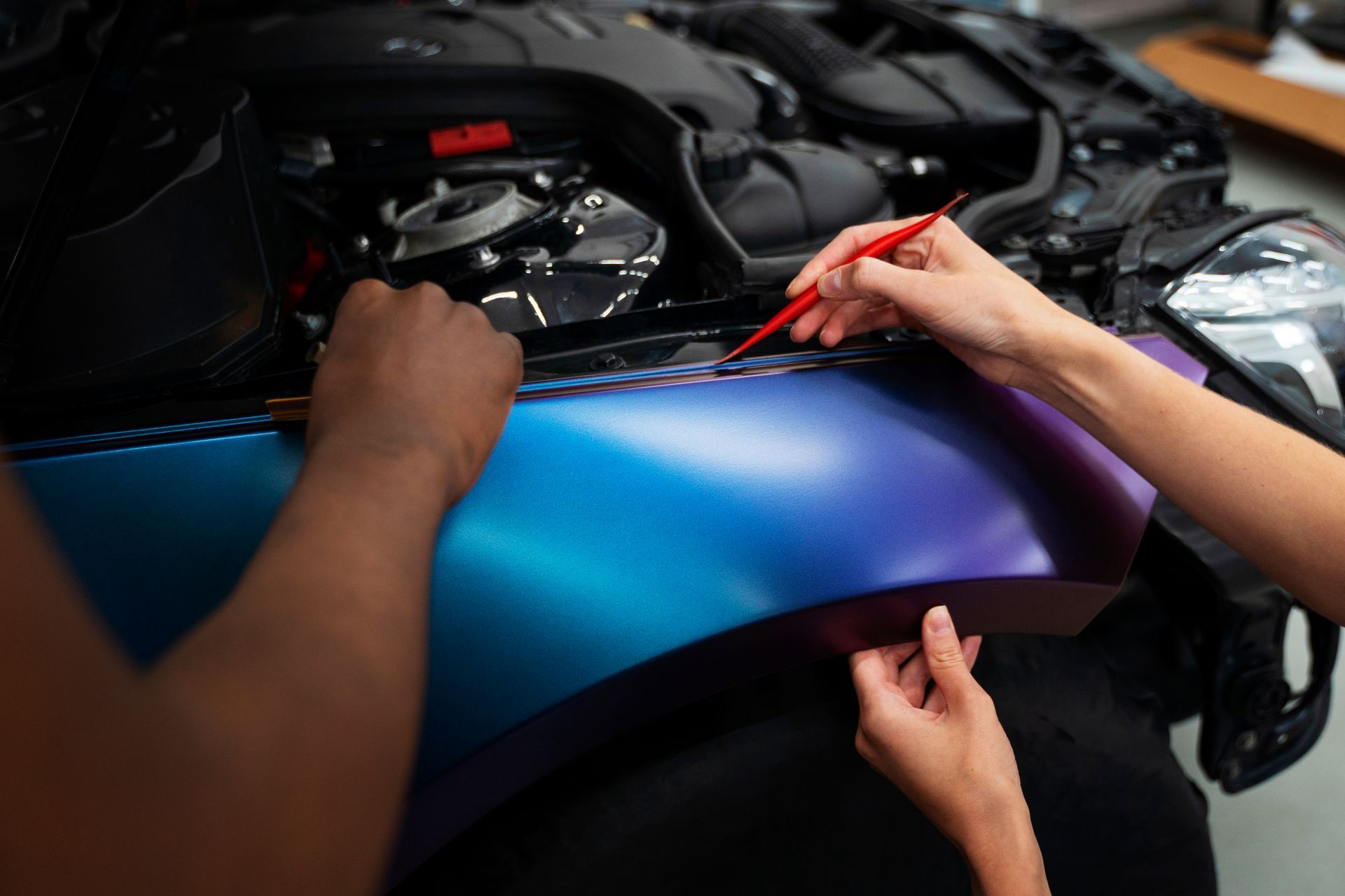 Hands applying blue-purple vinyl wrap to a car hood in an auto workshop