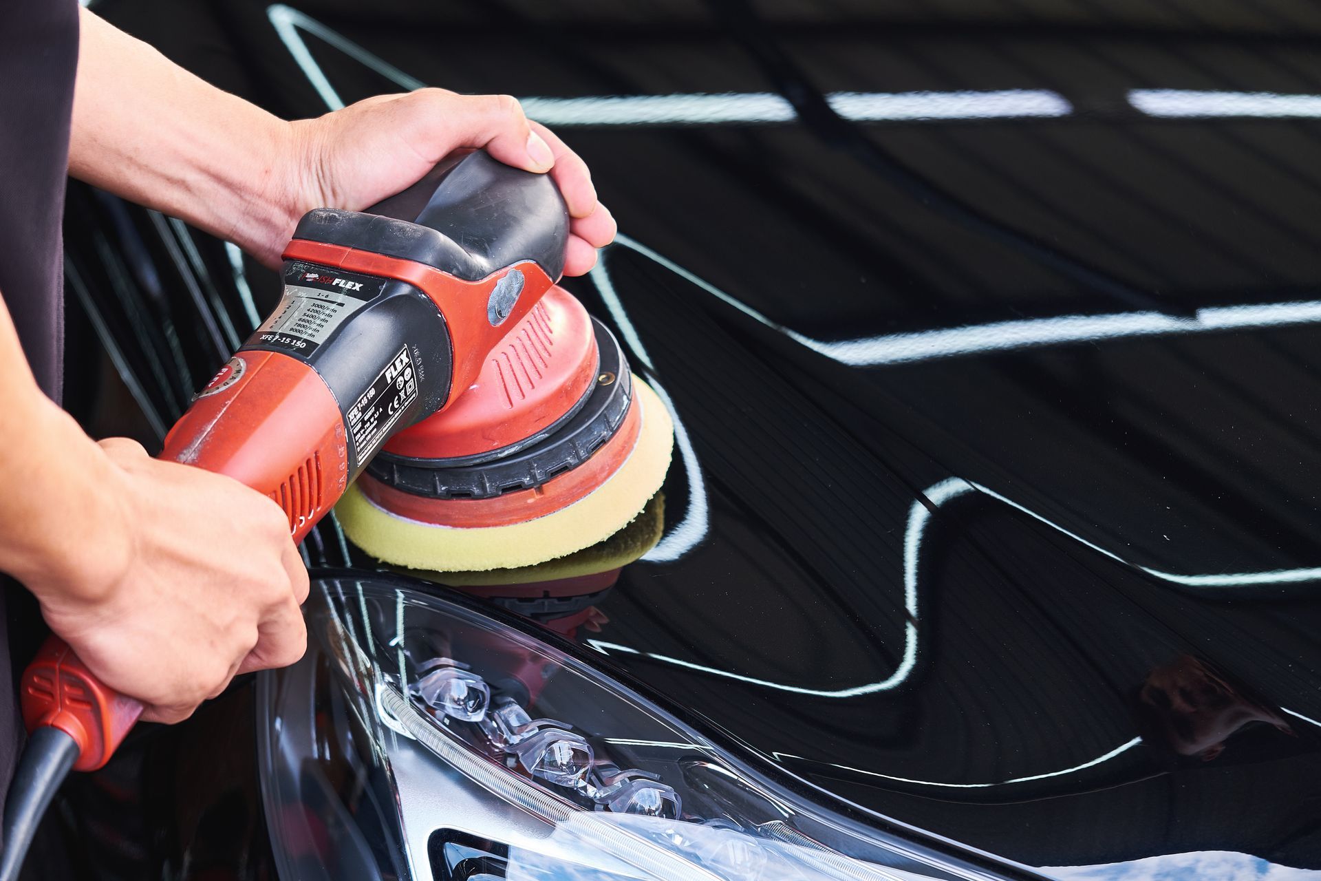 Hands polishing a black car with a red buffer in a garage