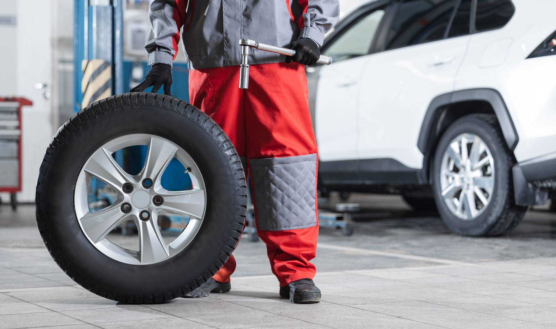 Mechanic in red uniform holding a tire beside a white car in a garage