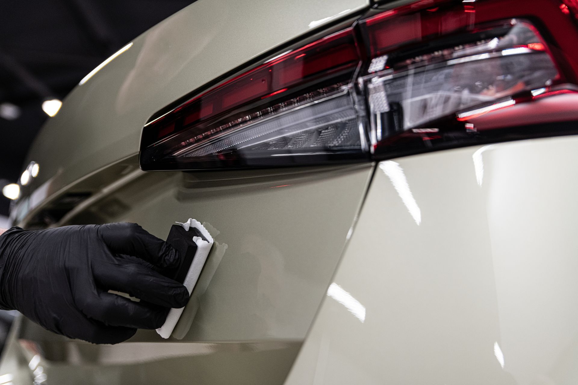Gloved hand polishing a beige car’s rear panel near the tail light in a garage