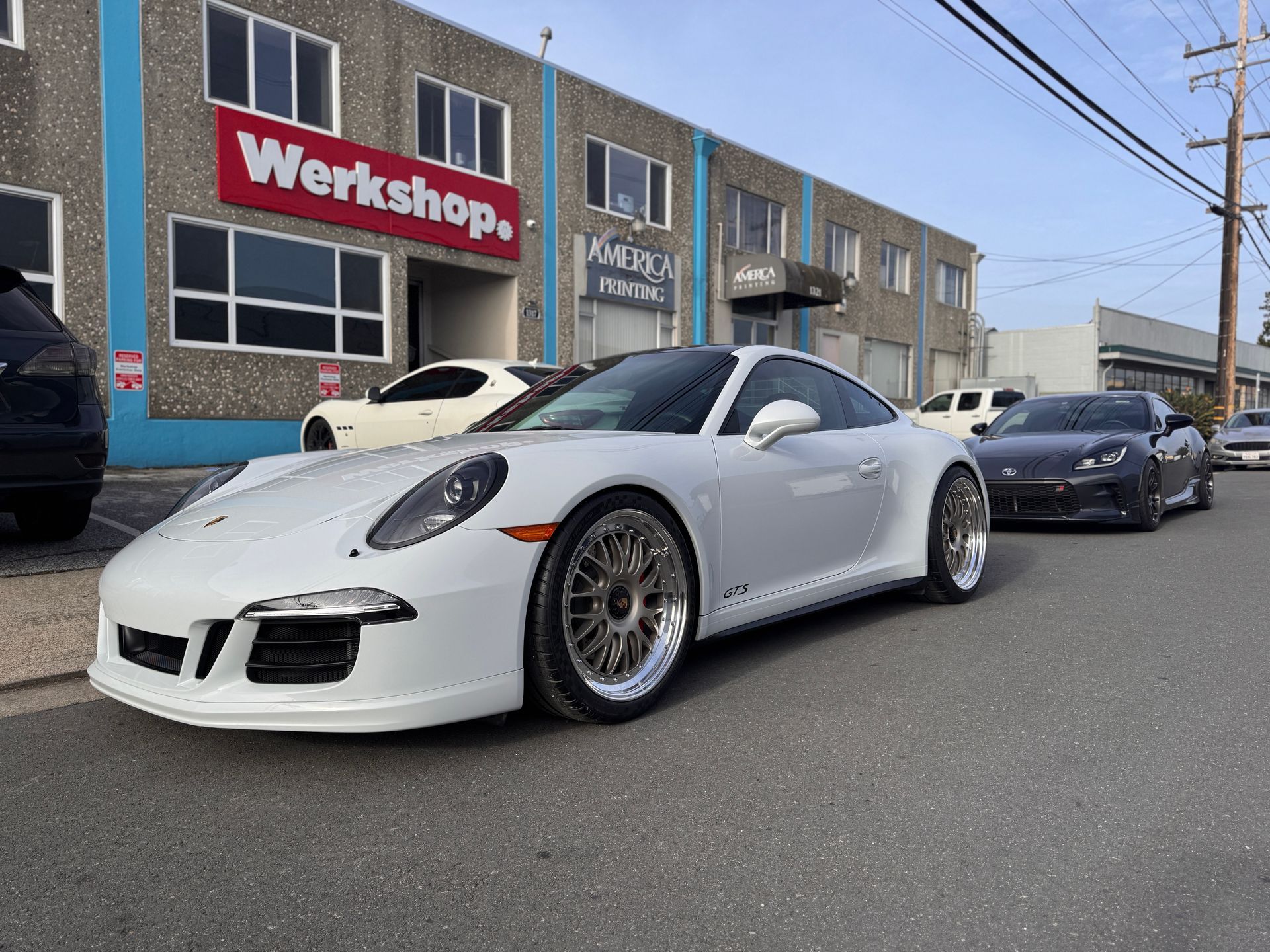 White sports car parked on a street outside a workshop, with another car behind it.