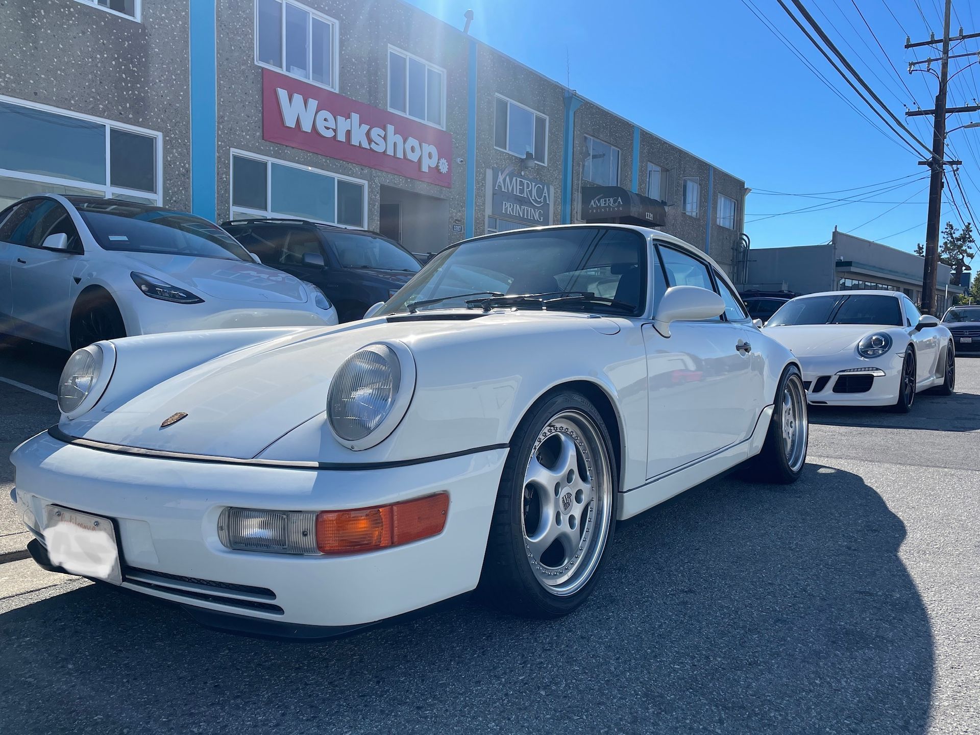 White Porsche 911 parked outside a workshop, with another white car behind it on a sunny street.