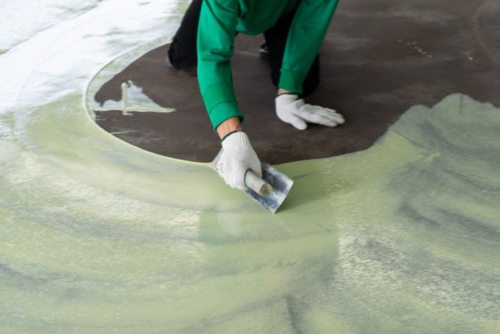 Person in green overalls applying material to a concrete floor with a hand tool.