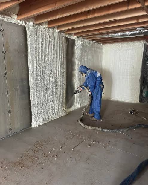 Worker in blue spray suit applying insulation to a basement wall.