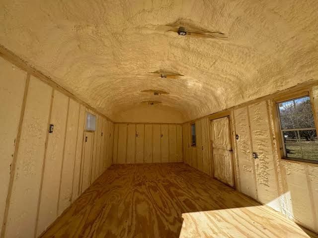 Empty unfinished room with plywood walls, arched insulated ceiling, and a window on the right.