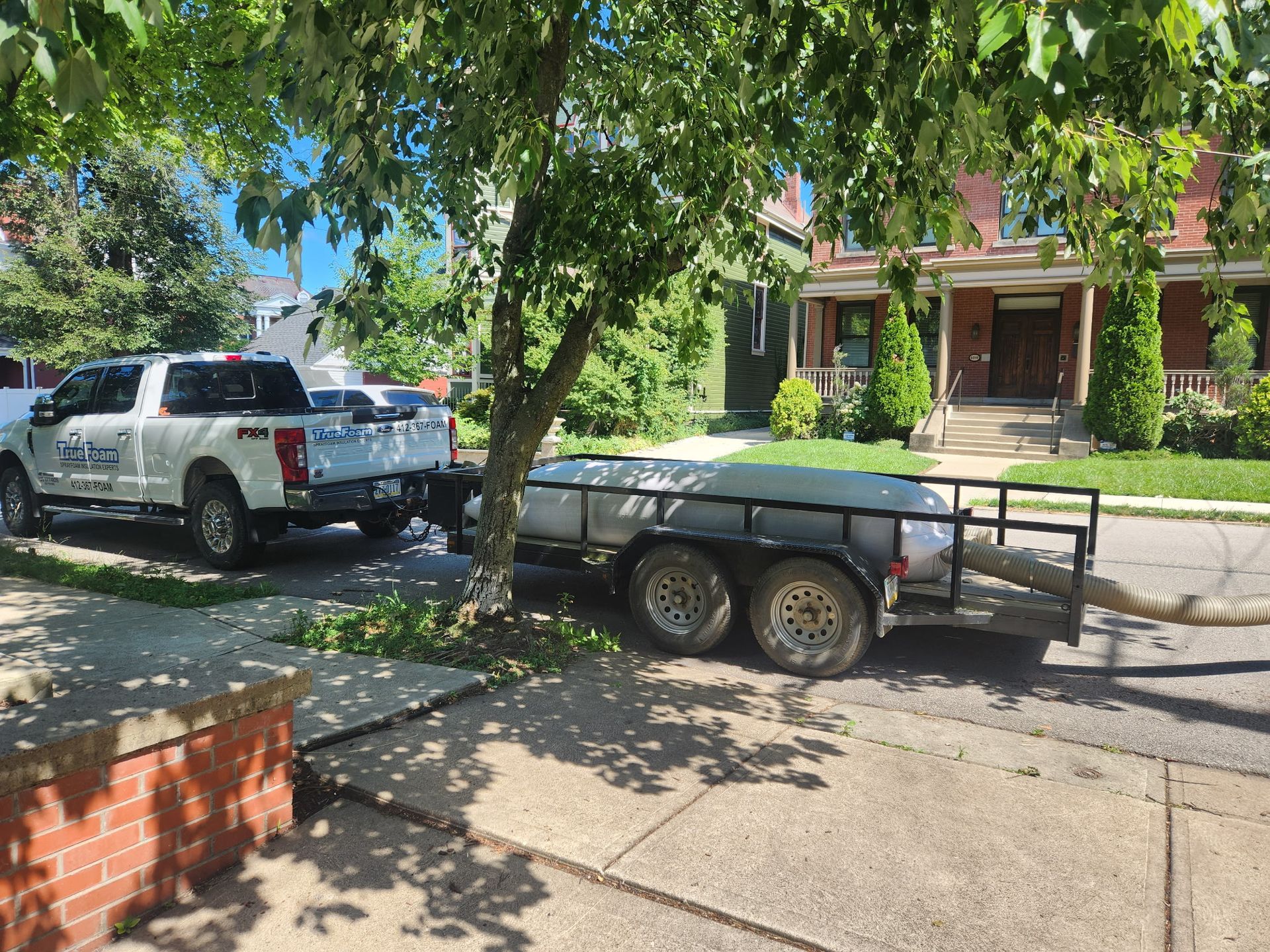 White pickup truck towing a gray trailer parked on a leafy suburban street in front of houses.