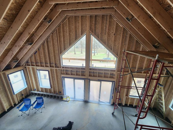 Unfinished attic room with exposed wood framing, windows, ladders, and blue chairs on a concrete floor.