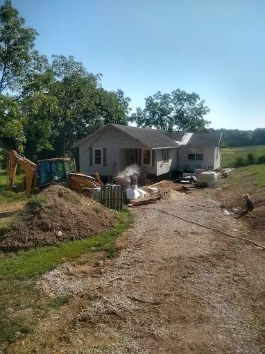 Small house under construction with dirt driveway, excavator, building materials, and trees in a rural yard