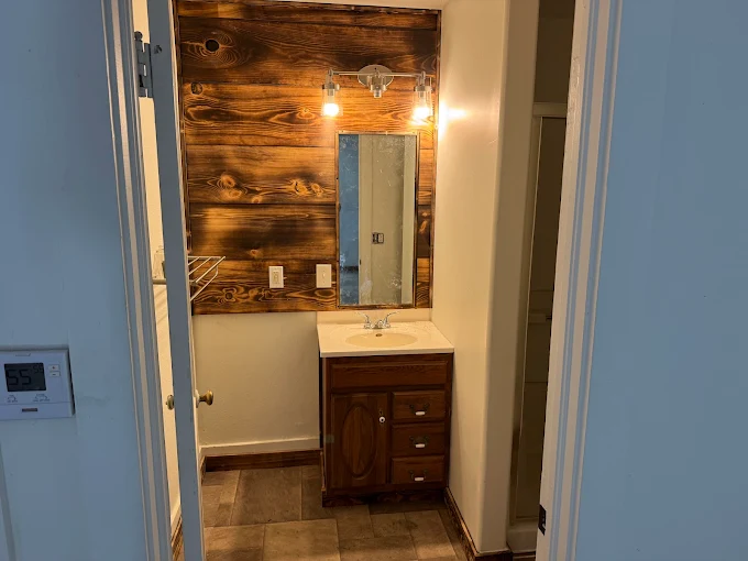 Small bathroom vanity with mirror, wood accent wall, and warm lighting seen through a doorway