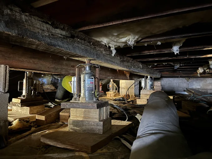 Crawlspace under a house with pipes, wooden beams, and a worker inspecting a floor jack under the floor
