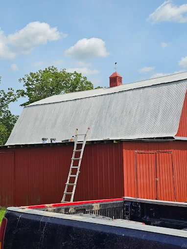 Red barn with gray metal roof, ladder leaning against it, under a blue sky with clouds.