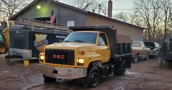 Yellow GMC dump truck in a muddy yard beside a garage at dusk