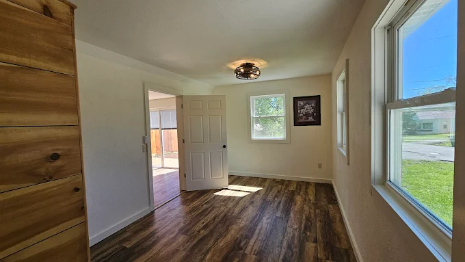 Bright empty room with wood floors, white walls, open door, and large windows overlooking a lawn