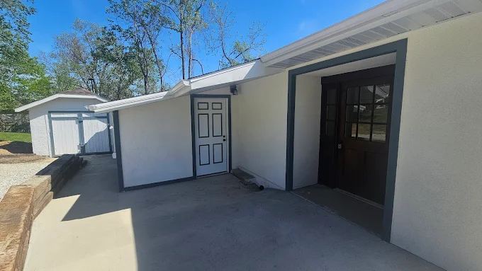 Side yard with a concrete patio, white stucco house, and open black doorway beside a white door.