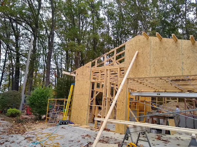 Wood-framed house under construction beside a wooded area, with exposed sheathing and scaffolding.