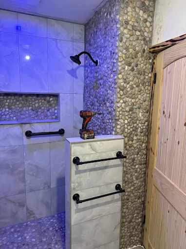 Small bathroom shower with white tile, blue light, pebble accent wall, and a wooden door.