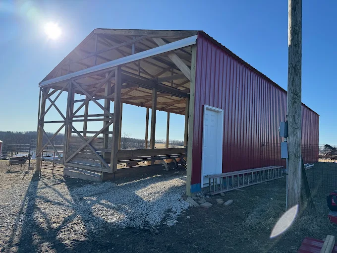 Partially built red barn or shed with open framing on a gravel lot in bright sunlight