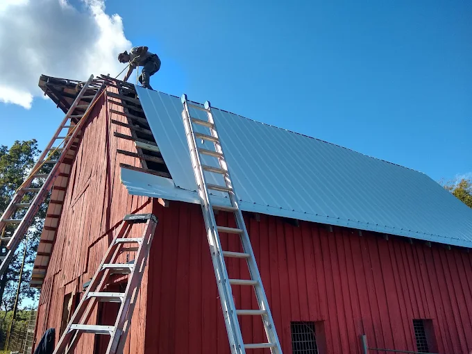 Two ladders leaning against a red barn roof under blue sky, with a worker at the peak.
