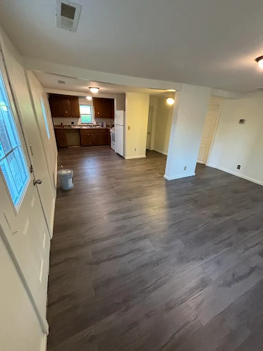 Empty open-plan living room with wood floors, white walls, and a kitchen in the background.