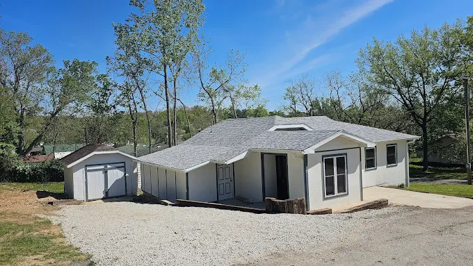 Small beige house with attached garage on a gravel lot under a clear blue sky