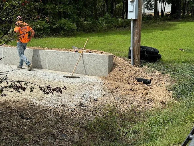 Worker in orange vest clearing debris beside a concrete barrier and utility pole on a grassy roadside.