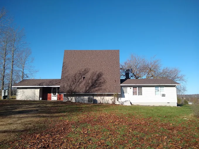 Rural house with a large brown roof, white wings, and a leaf-covered yard under a clear blue sky