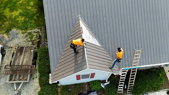 Two workers on a gray roof, one using a ladder at the edge of a house.