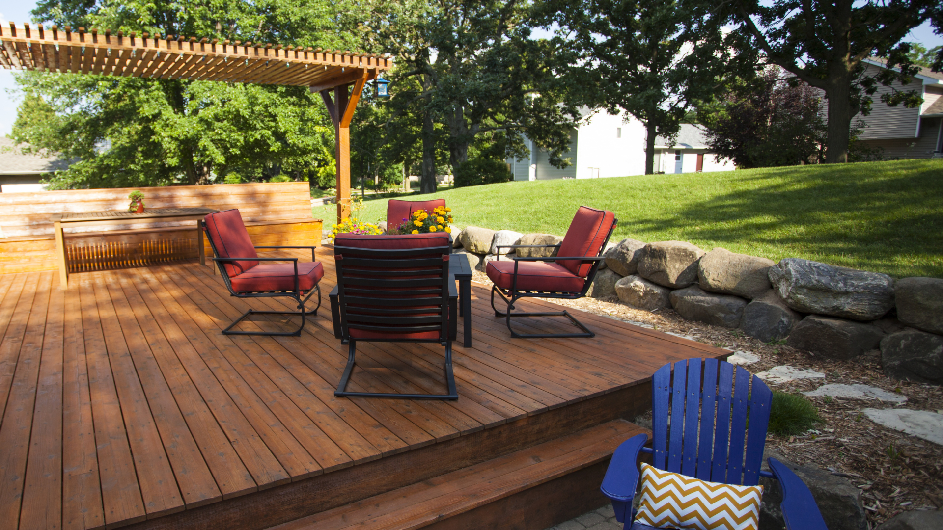 Wooden deck with red patio chairs, pergola, and stone retaining wall in a sunny backyard
