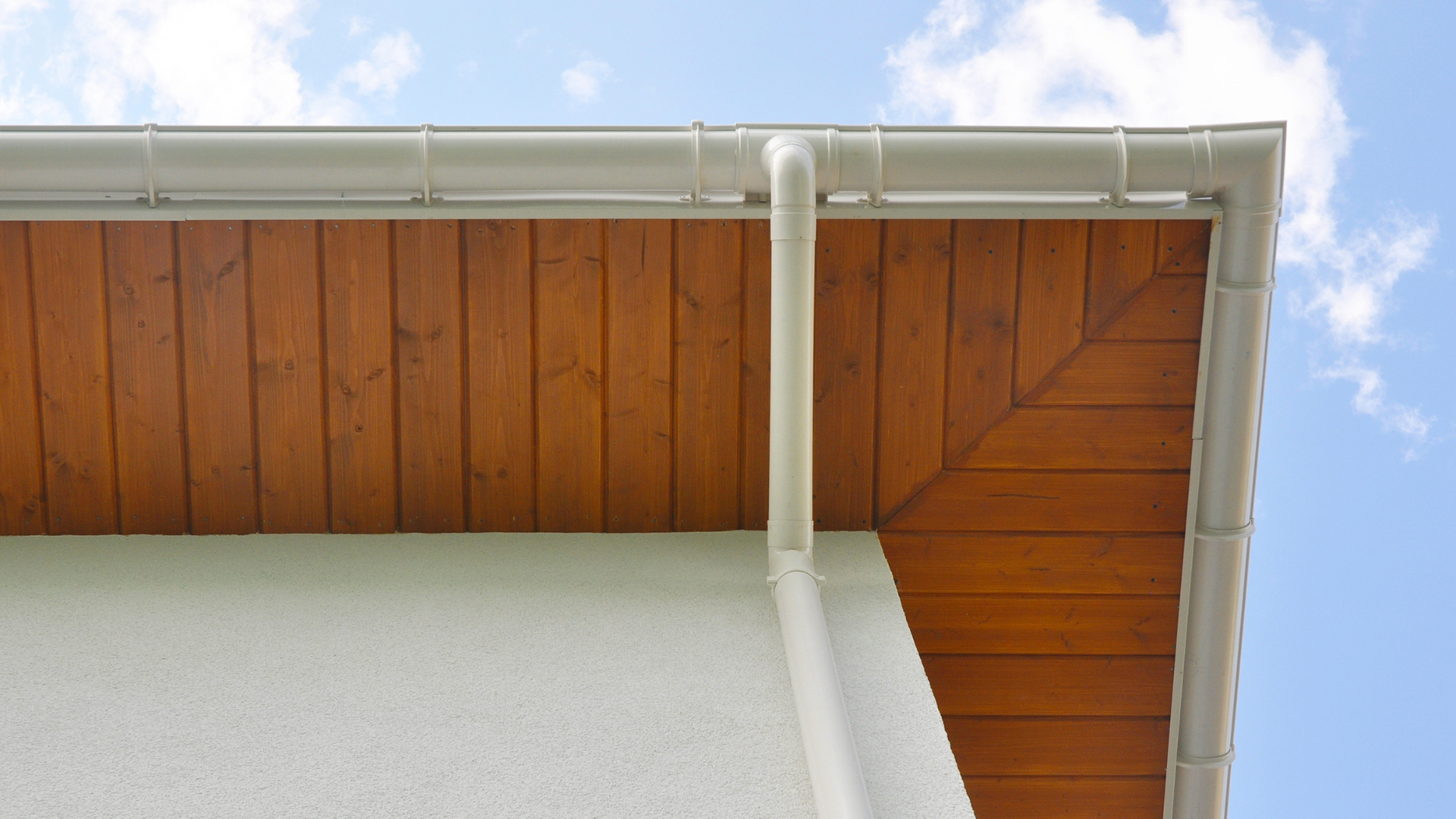 White house eave with gutter and downspout, showing a wooden soffit against blue sky