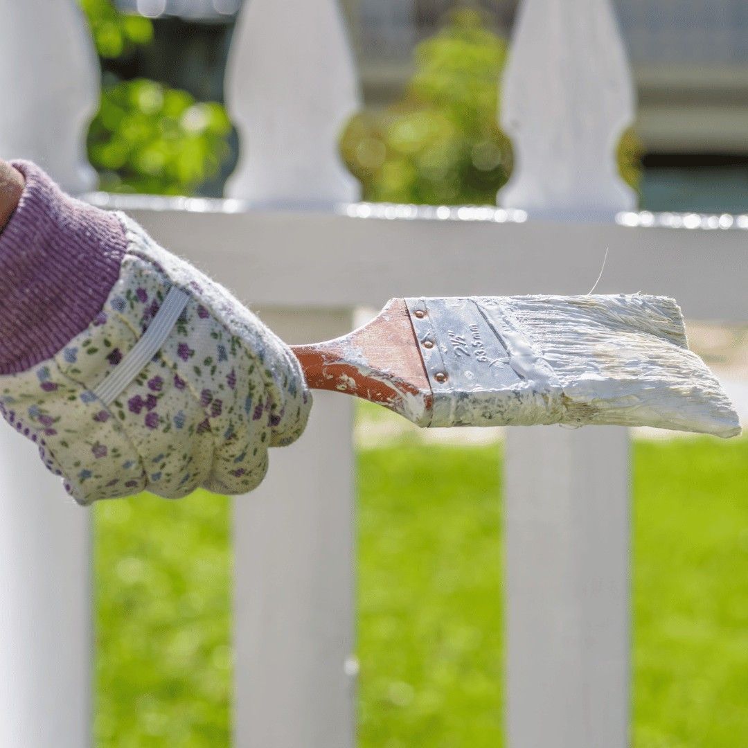Gloved hand painting a white fence with a brush in a sunny yard
