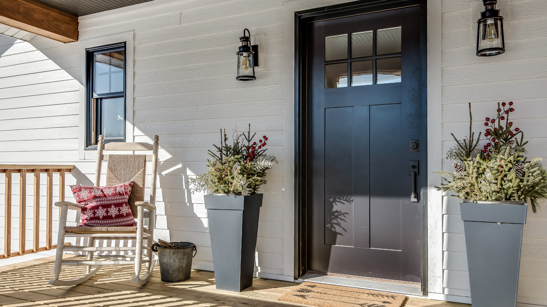 Front porch with black door, white brick walls, lanterns, potted flowers, and a wooden chair with cushion