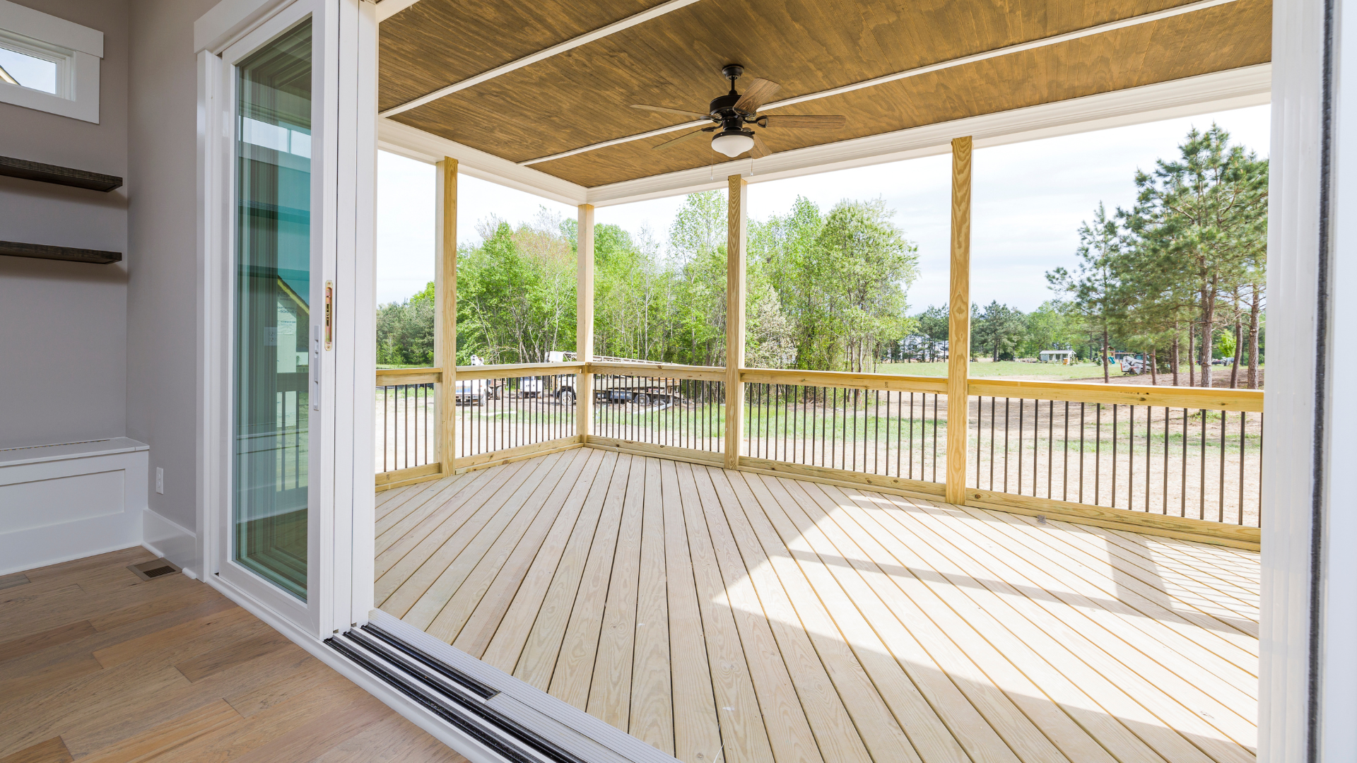 Covered porch with light wood decking, railing, and ceiling fan overlooking a grassy yard and trees