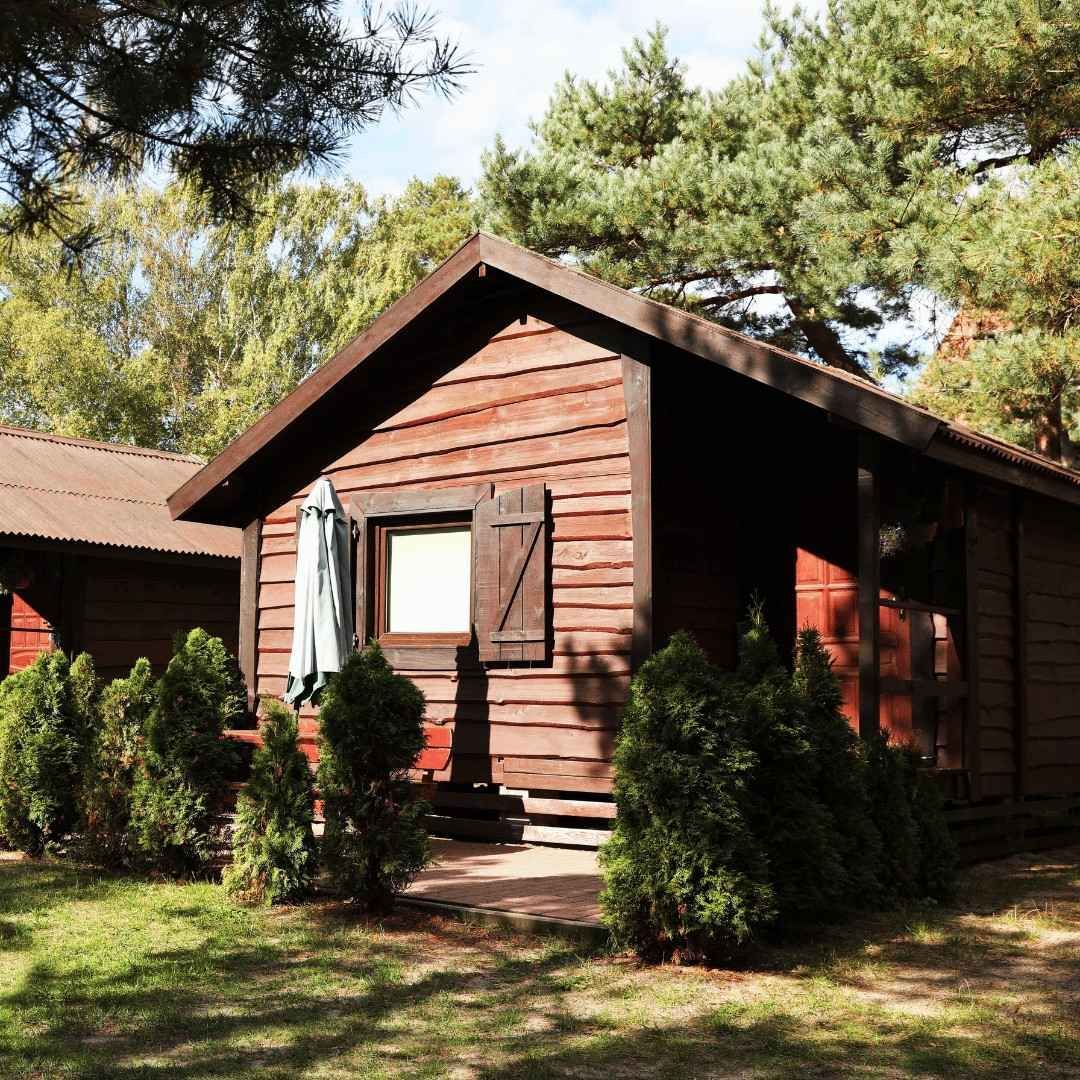 Wooden cabin with red-brown siding and a small porch, surrounded by evergreen trees in a sunny yard.