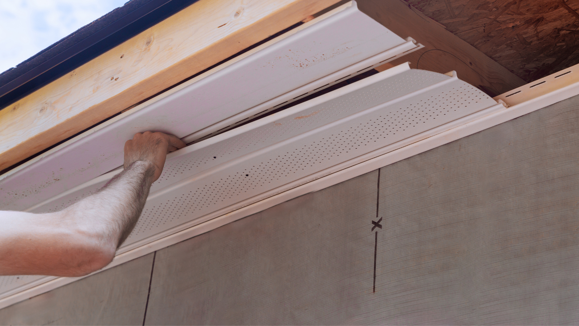 Underside of a roof eave with exposed wooden rafters and white soffit panels under a beige wall