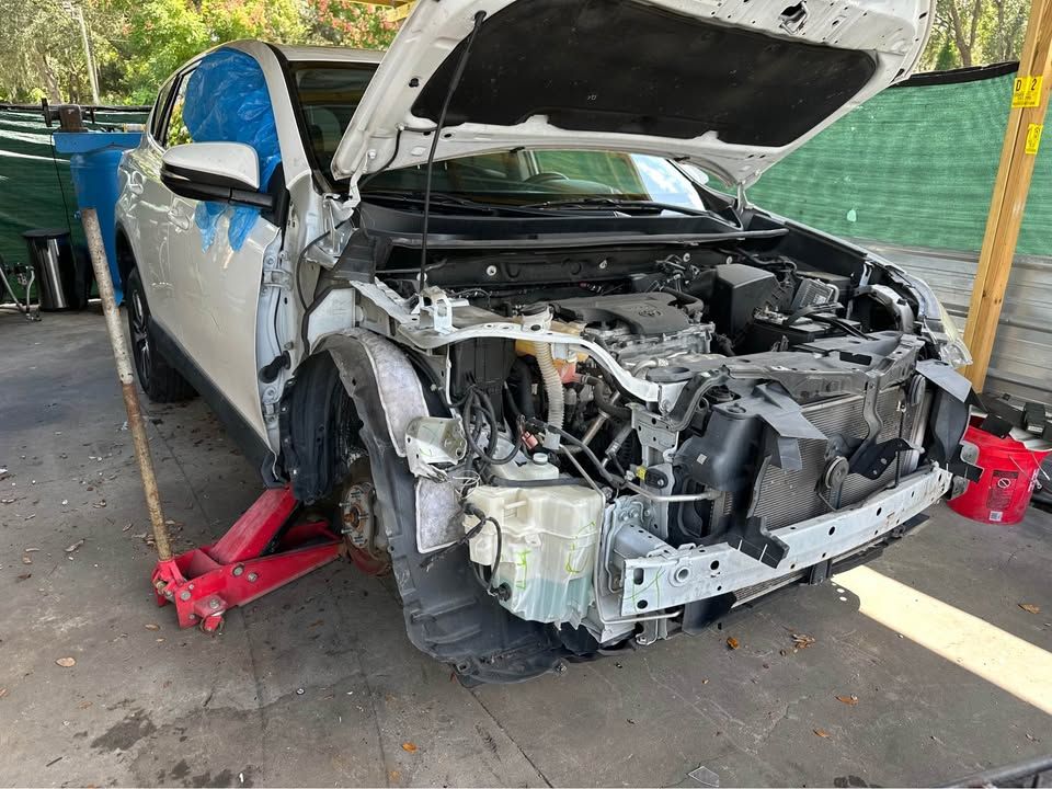White car with front-end damage, hood open, supported by a red jack, outdoors.