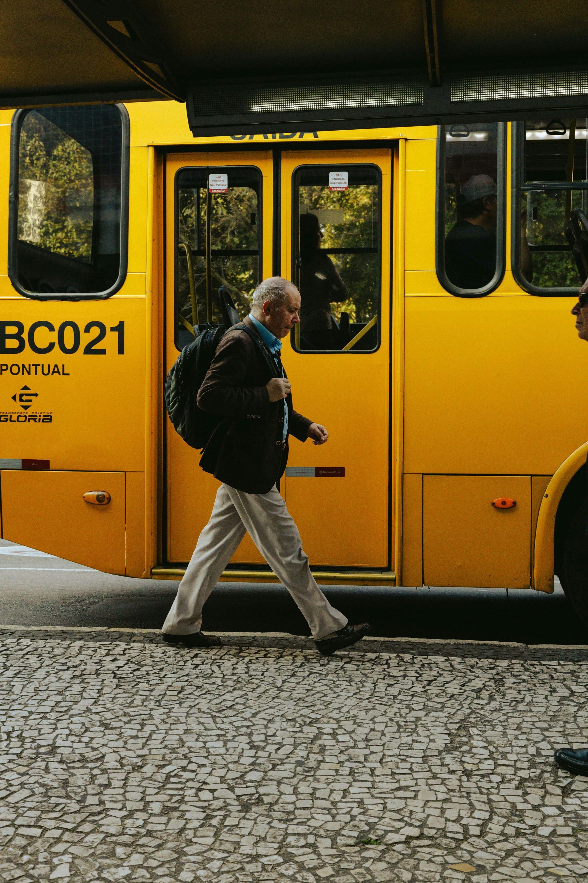 Older man with backpack walks past yellow bus on cobblestone street.