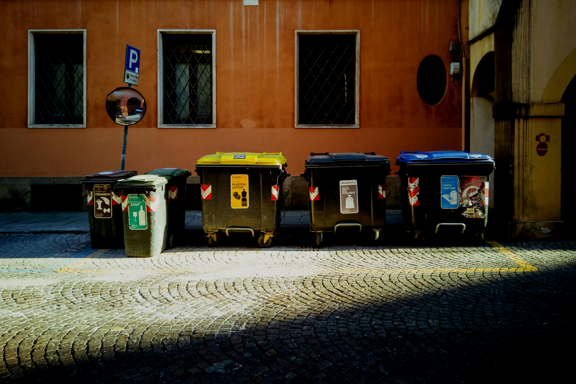 Row of colorful recycling bins on cobblestone street, orange building backdrop.