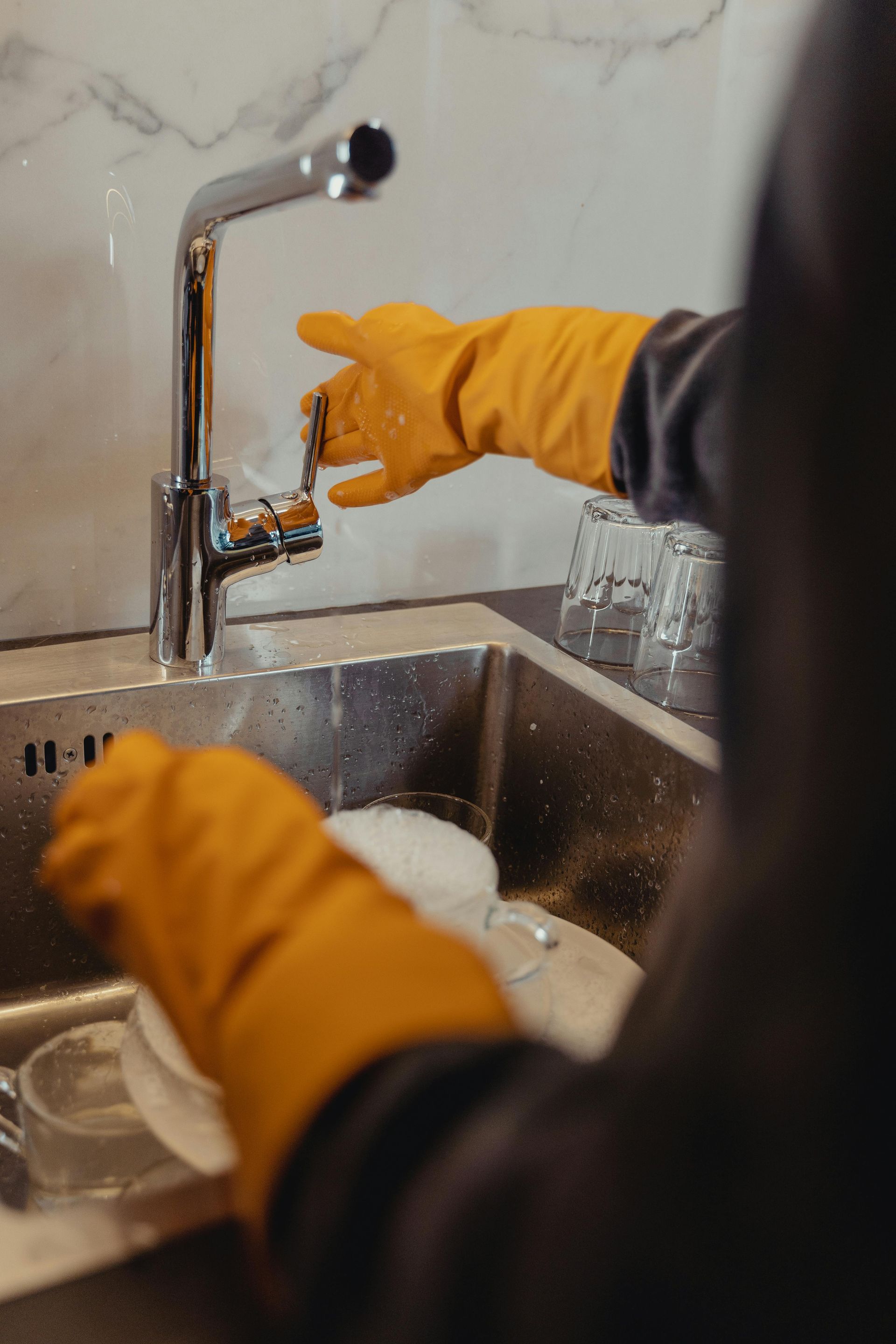 Person wearing orange gloves washes dishes in a stainless steel sink under a chrome faucet.